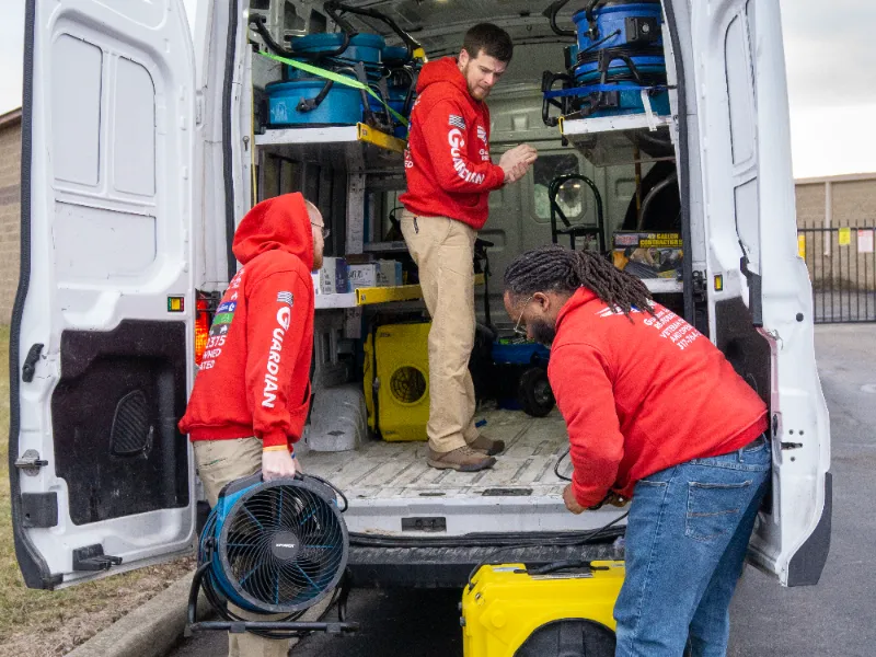 Three Guardian Angel Restoration crew members in red branded hoodies loading commercial water mitigation equipment into the open rear doors of a service van, ready for commercial property response