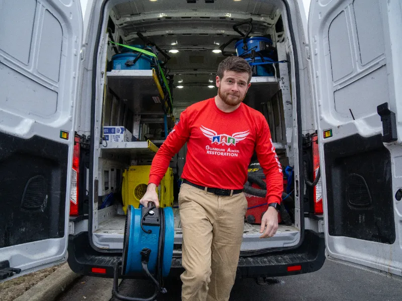 Guardian Angel Restoration technician in red shirt carrying blue commercial drying equipment out of a service van toward a damaged Noblesville home