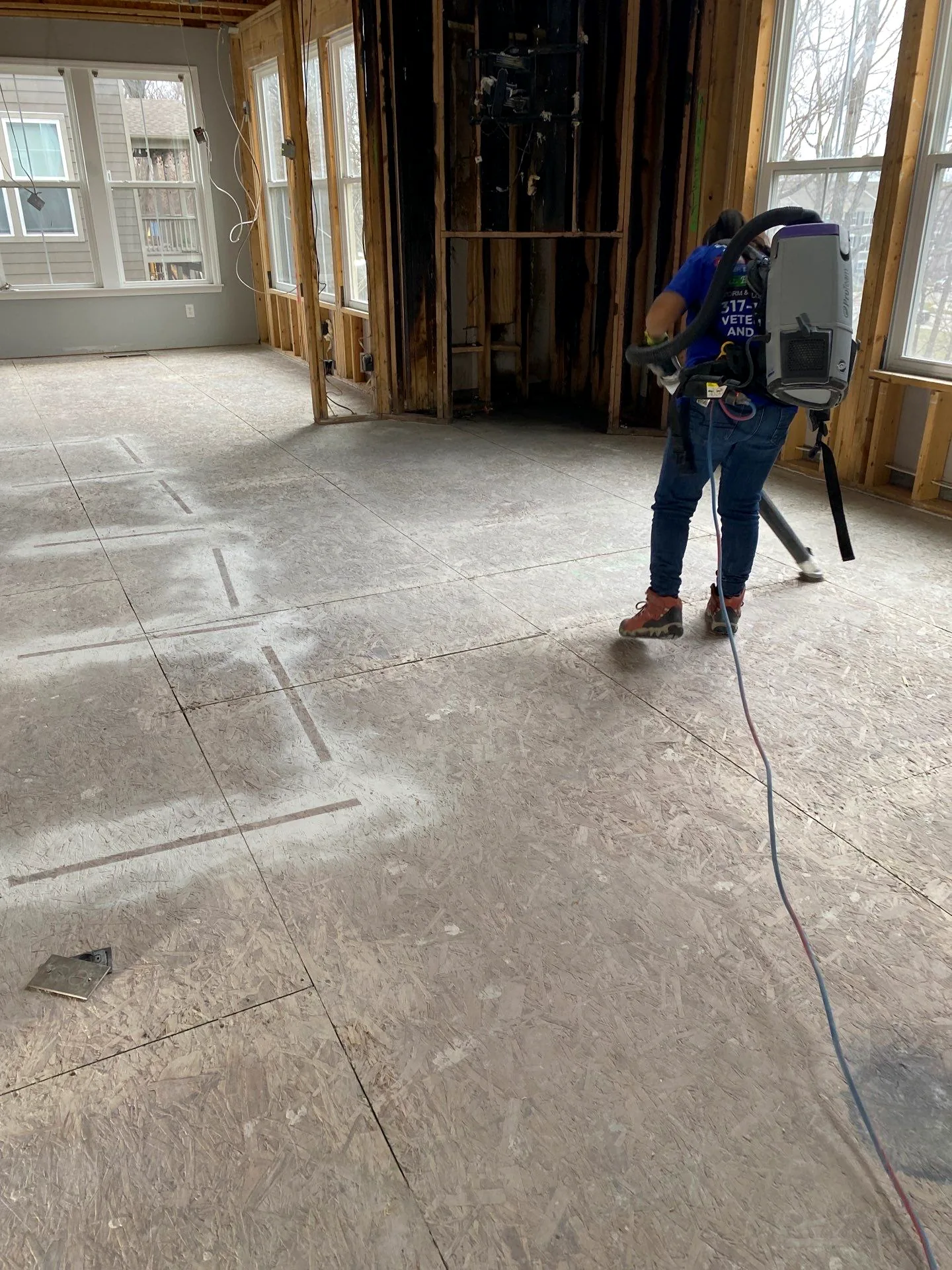 Technician using a backpack HEPA vacuum on new subfloor beside a charred wall during fire cleanup.