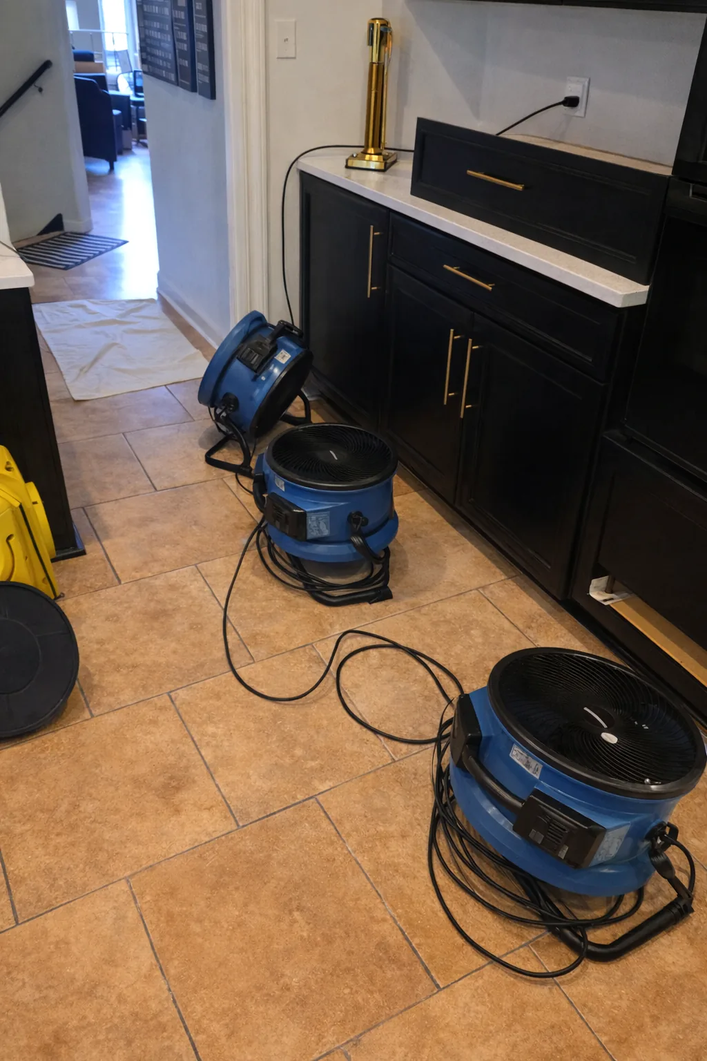 Air movers drying a kitchen tile floor beside dark cabinets after water damage.