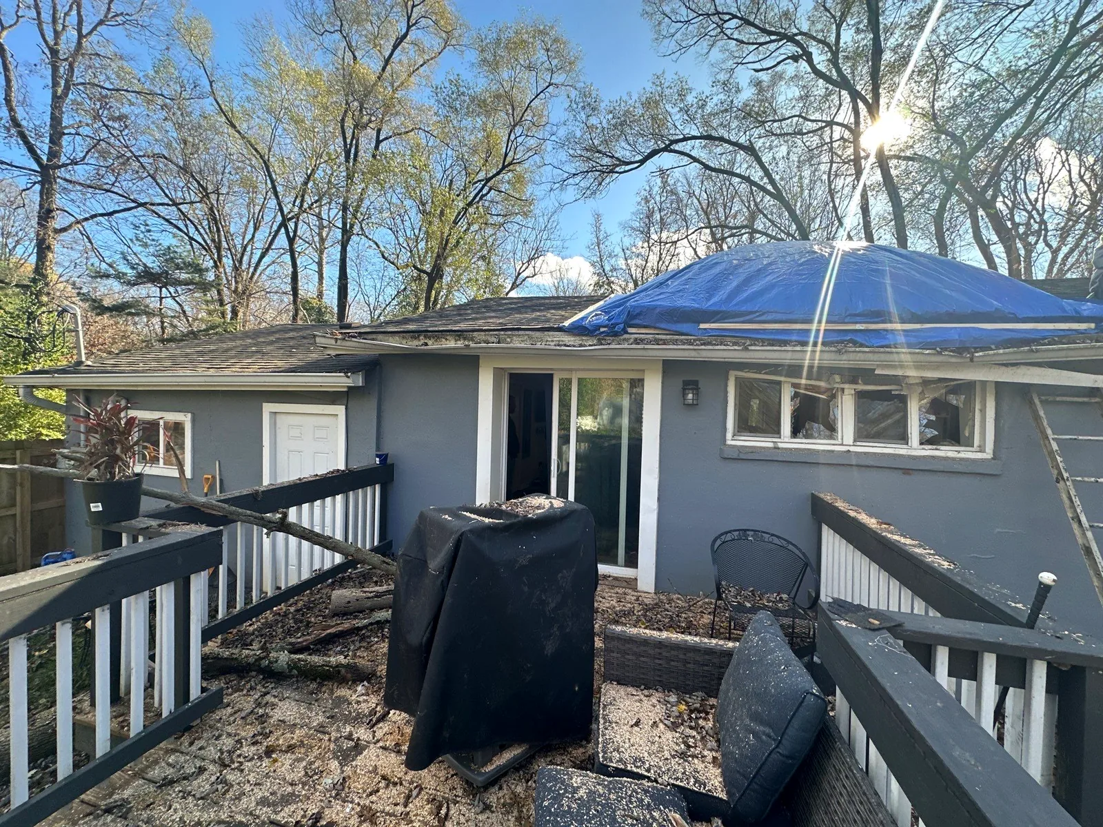 Blue emergency tarp covering the roof of a home after a fallen tree, with debris across the deck.