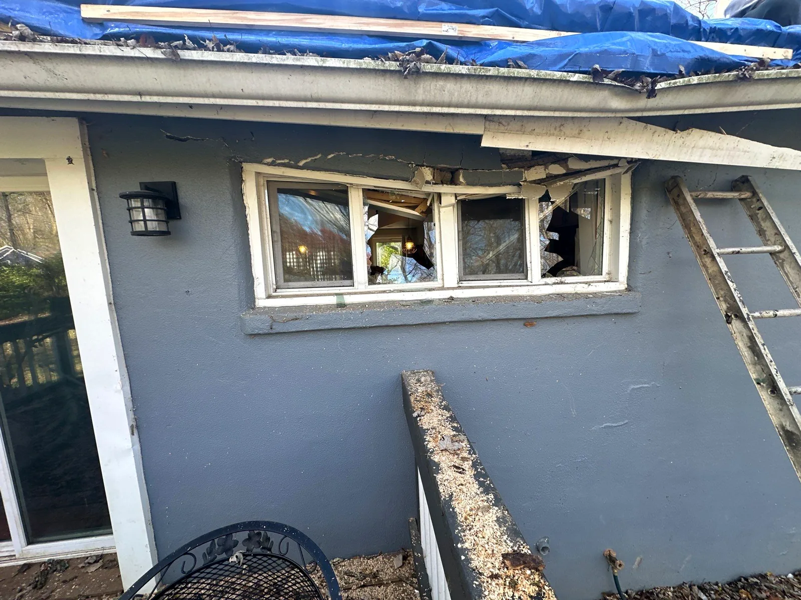 Broken windows and a crushed eave on the rear of a home after a tree strike, with a tarped roof above.