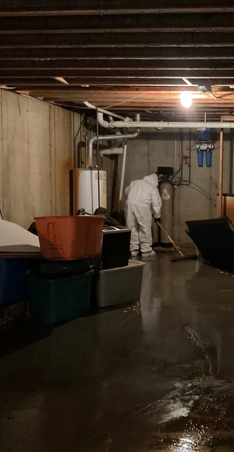Technician in a protective suit squeegeeing standing water across a flooded basement floor.