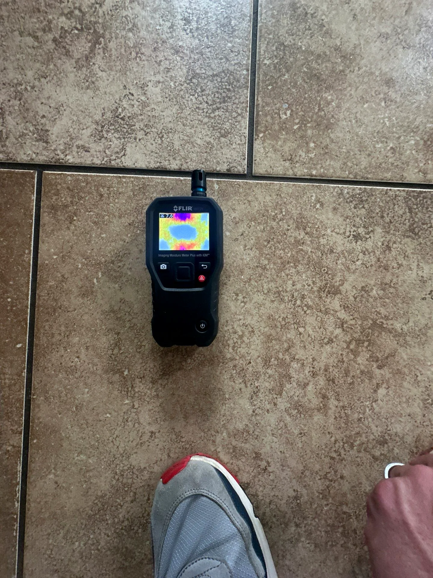 Thermal imaging moisture meter on a tile floor showing a wet spot on its screen during moisture mapping.