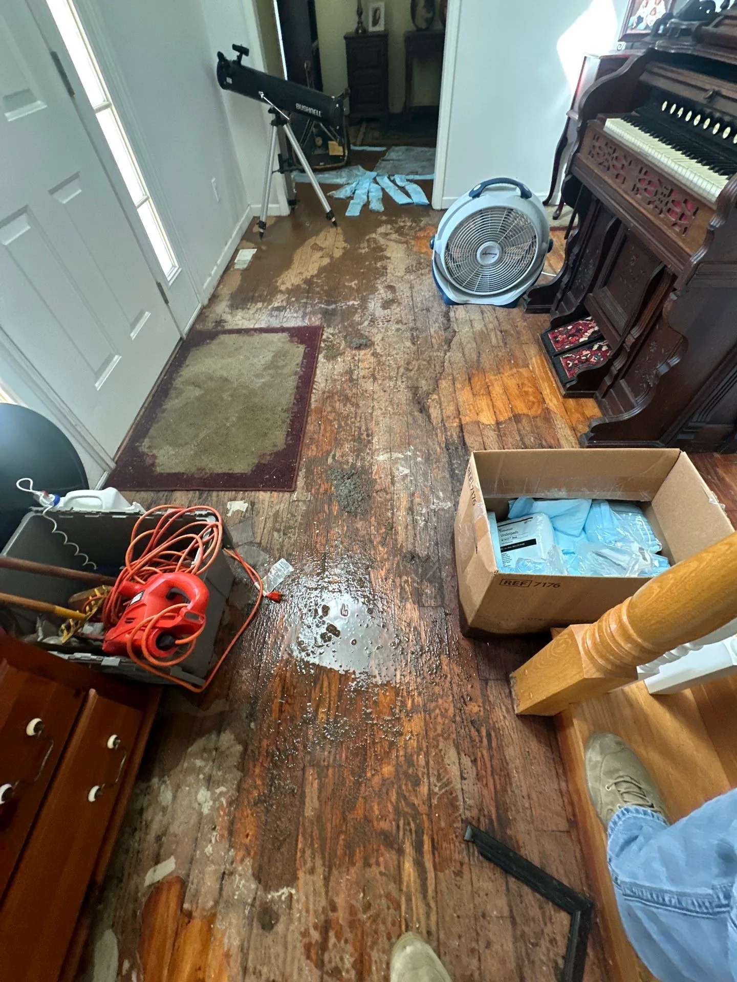 Wet hardwood entry with a fan running and belongings moved aside after water intrusion.